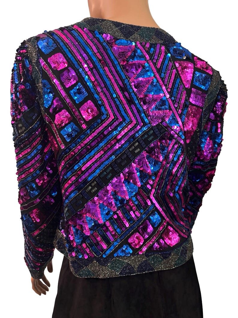 Puede incluir: Una chaqueta deslumbrante con un patr&oacute;n geom&eacute;trico de lentejuelas en tonos azules, morados y rosas. La chaqueta tiene mangas largas y una base negra, con un cuello y dobladillo brillantes. El dise&ntilde;o es intrincado y llamativo.