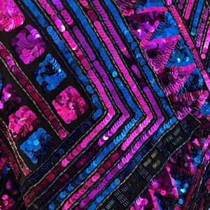 Puede incluir: Primer plano de una prenda con fondo negro, adornada con lentejuelas vibrantes en tonos azules, rosas y morados. Las lentejuelas est&aacute;n dispuestas en patrones geom&eacute;tricos, creando una superficie texturizada y brillante. El dise&ntilde;o presenta l&iacute;neas diagonales y formas cuadradas.