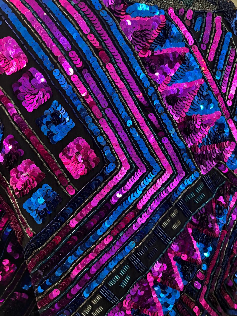 Puede incluir: Primer plano de una prenda con fondo negro, adornada con lentejuelas vibrantes en tonos azules, rosas y morados. Las lentejuelas est&aacute;n dispuestas en patrones geom&eacute;tricos, creando una superficie texturizada y brillante. El dise&ntilde;o presenta l&iacute;neas diagonales y formas cuadradas.