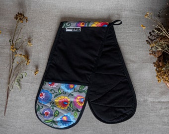Puede incluir: Manopla doble para horno negra con detalles florales. La manopla presenta un fondo gris con flores coloridas en tonos azules, rosas y amarillos. Tiene un pequeño lazo para colgar.