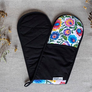 May include: Black double oven mitt with a floral pattern in white, blue, pink, and yellow. The mitt has a quilted black interior and a loop for hanging. The floral pattern is on the top side of the mitt.