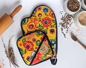 Puede incluir: Juego de manopla y agarradera amarillas con un estampado floral en rojo, azul y verde. El juego tiene ribetes negros y una etiqueta que dice "KOMPLET KUCHENNY". Un rodillo de madera y otros artículos de cocina están en el fondo.