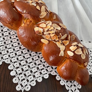 May include: A golden-brown, braided loaf of bread, topped with sliced almonds. The bread sits on a white, lace-edged cloth, creating a visually appealing contrast. The baked bread has a glossy finish, suggesting a rich, buttery texture. A delicious treat.