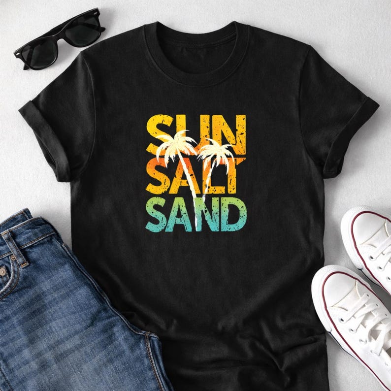 May include: Black t-shirt with the words "SUN SALT SAND" in a distressed, colorful font, with palm tree graphics. Sunglasses, blue jeans, and white sneakers are also visible.