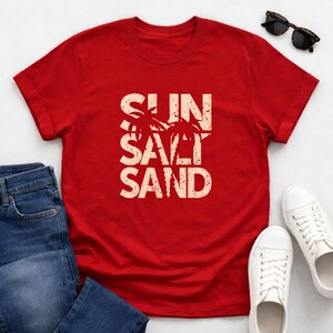 May include: A red t-shirt with the words "SUN SALT SAND" in a distressed, off-white font. Two palm trees are featured in the design. The shirt is paired with blue jeans, white sneakers, and sunglasses.