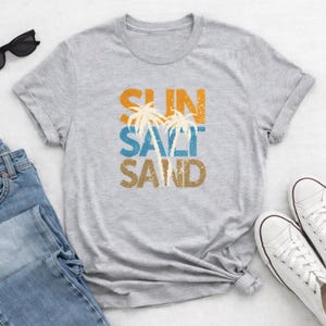 May include: A gray t-shirt with the words "SUN SALT SAND" in orange, blue, and brown, with palm tree graphics. The shirt is folded, with sunglasses, jeans, and white sneakers in the background.