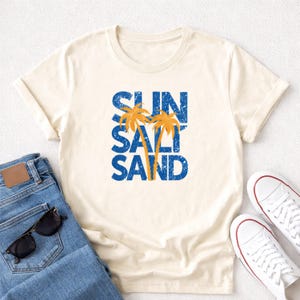 May include: Cream-colored t-shirt with the words "SUN SALT SAND" in blue, with two orange palm trees. The shirt is paired with blue jeans, sunglasses, and white sneakers.