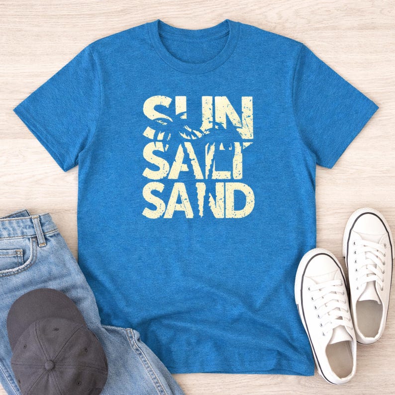 May include: A blue t-shirt with the words "SUN SALT SAND" in a distressed, off-white font. The design includes palm tree silhouettes. The shirt is paired with blue jeans, a gray cap, and white sneakers.