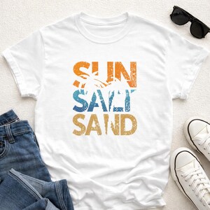 May include: White t-shirt with the words "SUN SALT SAND" in orange, blue, and tan block letters. A palm tree silhouette is visible within the word "SUN". The shirt is paired with blue jeans, white sneakers, and black sunglasses.