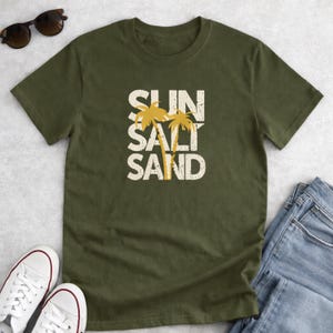 May include: Olive green t-shirt with the words "SUN SALT SAND" in a distressed white font, with two yellow palm trees. The shirt is paired with white sneakers, sunglasses, and blue jeans.