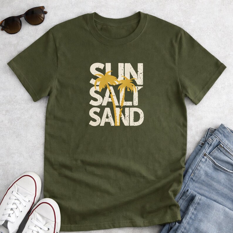 May include: Olive green t-shirt with the words "SUN SALT SAND" in a distressed white font, with two yellow palm trees. The shirt is paired with white sneakers, sunglasses, and blue jeans.