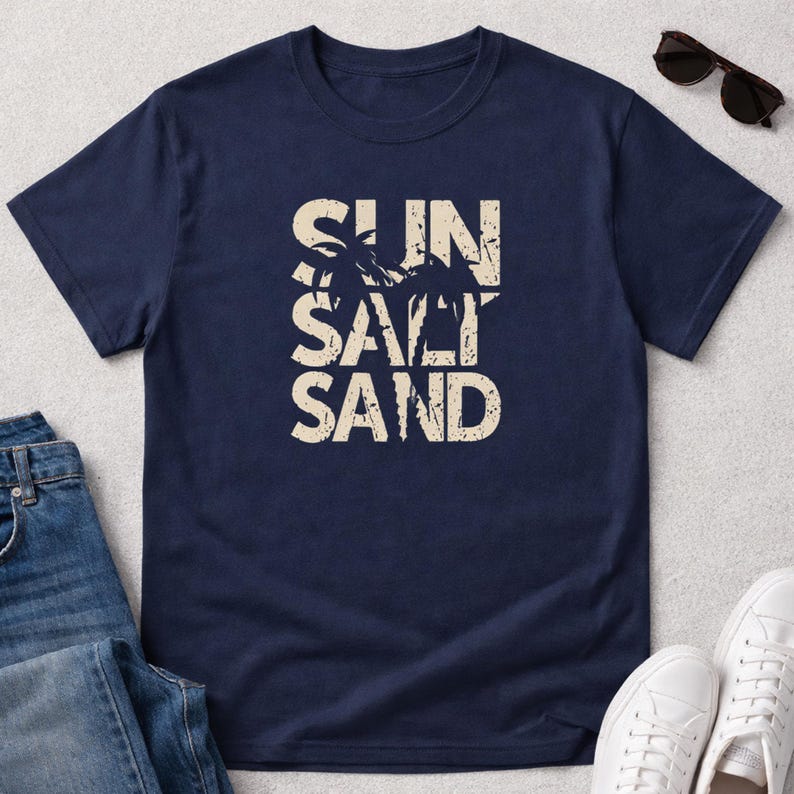 May include: Navy blue t-shirt with the words "SUN SALT SAND" in a distressed, off-white font. The design includes silhouettes of palm trees. Sunglasses, jeans, and white sneakers are also visible.