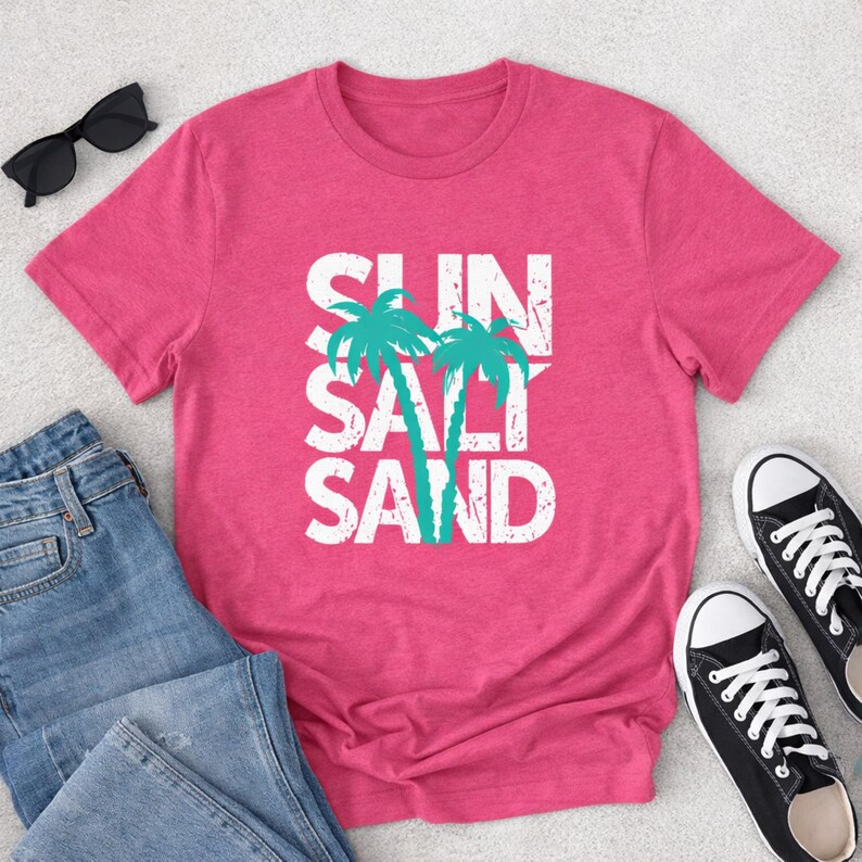 May include: A pink t-shirt with the words "SUN SALT SAND" in white block letters and two turquoise palm trees. The shirt is paired with blue jeans, black sunglasses, and black and white sneakers, suggesting a beach or summer theme.