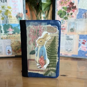 May include: A small, handmade journal with a blue cover and black spine. The front features a collage with a Peter Rabbit sticker, pink flowers, and a fern. The journal has a vintage aesthetic with torn paper and handwritten text.