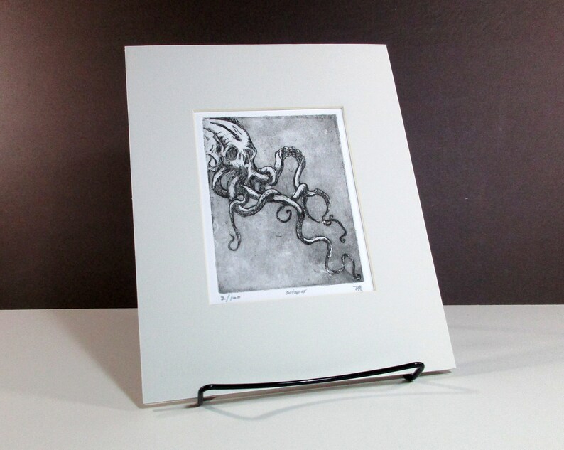 Puede incluir: Una impresi&oacute;n en blanco y negro de un pulpo con tent&aacute;culos detallados, enmarcada en un passe-partout blanco. La obra de arte se exhibe en un caballete de metal negro. La impresi&oacute;n est&aacute; numerada 2/100.