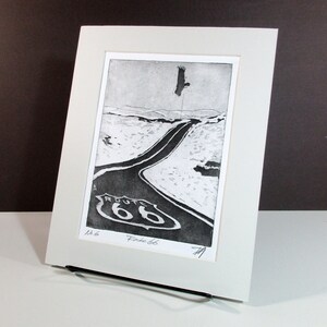 K&ouml;nnte beinhalten: Ein Schwarzwei&szlig;druck einer Stra&szlig;e mit einem Route 66-Schild. Die Stra&szlig;e ist kurvig und ein Vogel fliegt im Himmel dar&uuml;ber. Der Druck ist in einem wei&szlig;en Passepartout gerahmt.