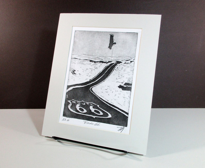 K&ouml;nnte beinhalten: Ein Schwarzwei&szlig;druck einer Stra&szlig;e mit einem Route 66-Schild. Die Stra&szlig;e ist kurvig und ein Vogel fliegt im Himmel dar&uuml;ber. Der Druck ist in einem wei&szlig;en Passepartout gerahmt.