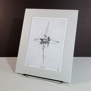 May include: A black and white etching of a sailboat with a single person on board. The sailboat is in the middle of the water with a reflection of the boat in the water. The etching is framed in a white mat and a white frame.
