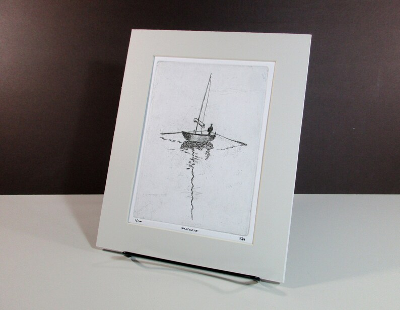 May include: A black and white etching of a sailboat with a single person on board. The sailboat is in the middle of the water with a reflection of the boat in the water. The etching is framed in a white mat and a white frame.