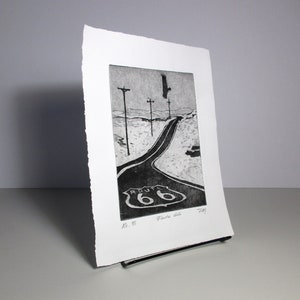 K&ouml;nnte beinhalten: Schwarzwei&szlig;druck einer Stra&szlig;e mit einem Route 66-Schild. Die Stra&szlig;e ist kurvig und es gibt Telefonmasten auf beiden Seiten. Ein Vogel fliegt im Himmel &uuml;ber der Stra&szlig;e.