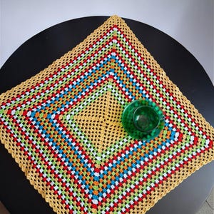Pode incluir: Uma toalha de mesa quadrada de crochê feita à mão com um design vibrante e colorido. O padrão apresenta quadrados concêntricos de vermelho, branco, azul, verde e laranja, emoldurados por uma borda amarela. Um pires de vidro verde está em cima.