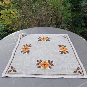 Op de afbeelding: Een vierkant beige linnen tafelkleedje met een kruissteekpatroon van vier oranje en gele bloemen met bruine bladeren. De stof heeft een zichtbare geweven textuur en is omrand met een dunne bruine lijn met kleine blaadjes in elke hoek.
