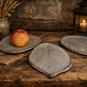 Può includere: Una natura morta rustica presenta tre piatti in ceramica a forma di foglia, uno con una mela rossa e gialla. I piatti sono su una superficie di legno, con un panno di juta, un vaso di grano e una lanterna accesa sullo sfondo.