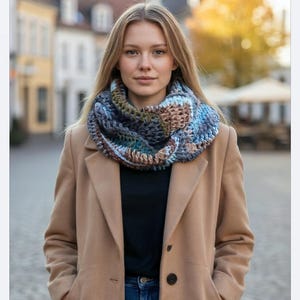 Puede incluir: Una mujer con un abrigo color canela y una bufanda infinita de ganchillo azul, marrón y verde. La bufanda es gruesa y parece hecha a mano. El fondo es una escena exterior borrosa con edificios y árboles.