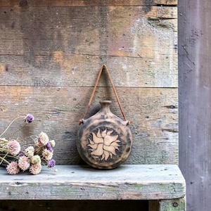 Op de afbeelding: Een ronde, bruine keramische fles met een zonnebloempatroon, opgehangen aan een leren riem. De fles wordt gepresenteerd tegen een verweerde houten achtergrond. Gedroogde bloemen in paars en crème liggen op een houten plank.
