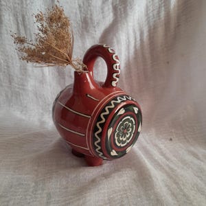 Op de afbeelding: Een rode keramische kan met witte en groene beschilderde ontwerpen. De kan heeft een rond lichaam en een klein handvat. Het is versierd met een bloemenpatroon en een golvende lijn.