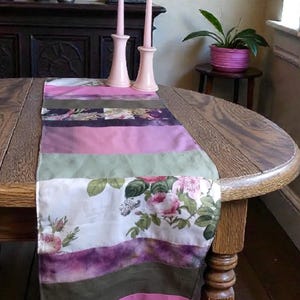 Può includere: Una colorata tovaglia con un design floreale e strisce rosa, verdi e viola. La tovaglia è posizionata su un tavolo di legno, con due candelabri rosa e candele sullo sfondo. Un pezzo decorativo per la sala da pranzo.
