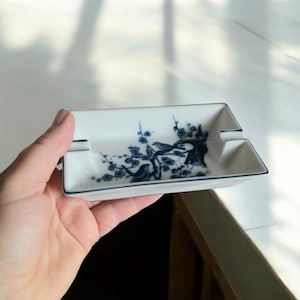 May include: White rectangular ashtray with a blue floral and bird design. The ashtray has a thin blue border and two grooves for resting cigarettes. The design features a tree branch with birds and flowers.