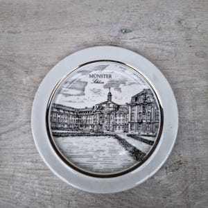 May include: A white ceramic plate with a silver rim and a black and white illustration of a building labeled "Münster Schloss". The plate is round and sits on a gray wooden surface.