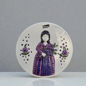 Puede incluir: Plato decorativo de cerámica blanca con una mujer con un vestido morado sosteniendo un ramo de flores moradas. El plato tiene un diseño en relieve con detalles florales morados y el texto "B. Johansson".