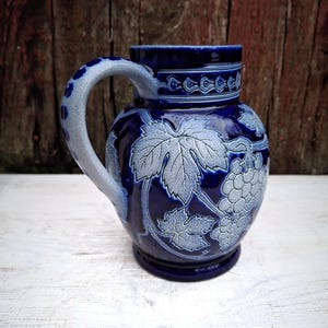 May include: A dark blue ceramic pitcher with a light blue handle and grape leaf design. The pitcher has a round body and a decorative band around the top. The handle is curved and has a dotted pattern.