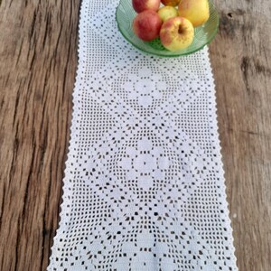May include: White crocheted table runner with a delicate pattern, laid on a wooden table with a bowl of red and yellow apples.