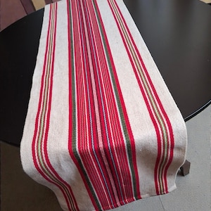 May include: A cream-colored table runner with vertical stripes in red, green, and blue. The runner is made of woven fabric and is draped over a black table. The design is simple and classic, suitable for various home decor styles.