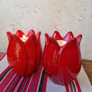 May include: Two glossy red ceramic candle holders shaped like tulip blossoms. Each holder features a scalloped rim and a smooth, rounded body. A small, lit white tea light candle is placed inside each of the two vibrant red tulip-shaped vessels.