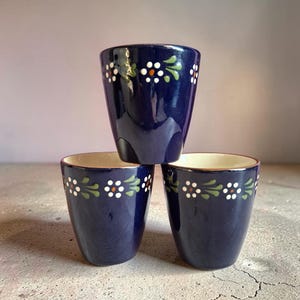 May include: Three navy blue ceramic cups stacked together. Each cup has a white, orange, and green floral pattern around the rim. The interior of the cups is cream-coloured.