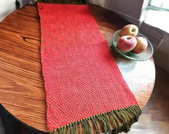 May include: Red table runner with a diamond pattern and green fringe. The runner is on a wooden table with a bowl of apples.