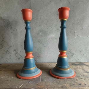 Può includere: Una coppia di portacandele in legno tornito con una silhouette a più livelli. I portacandele sono dipinti con fasce alternate di color corallo opaco, blu profondo e blu chiaro, con sottili anelli dorati che accentuano le sezioni centrali.
