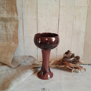 Puede incluir: Una copa de cerámica burdeos brillante, con un cuenco redondeado y un tallo delgado. La copa se exhibe sobre una superficie de madera clara. La altura de la copa es de unos 18 cm. Flores secas y una tela de arpillera en el fondo.
