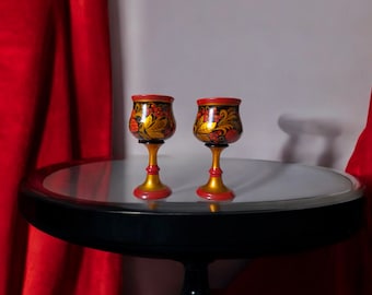 May include: Two hand-painted wooden shot glasses with intricate floral designs in red, yellow, and black on a black table with a reflective surface.