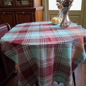 Può includere: Un tavolo rotondo coperto da una tovaglia a quadri rossi, turchese e bianchi. La tovaglia ha un bordo con frange. Un vaso di vetro trasparente con fiori secchi e due piccole zucche sono sul tavolo.