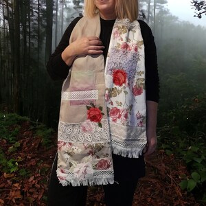Op de afbeelding: Een beige sjaal met bloemenpatronen en kantversiering. De sjaal is gemaakt van verschillende stoffen en heeft een patchwork-ontwerp. De sjaal wordt gedragen door een persoon in een bosrijke omgeving.