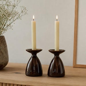 May include: Two dark brown, glossy ceramic candle holders with a flared, wide-rimmed top and a rounded, tapered base. Each holder contains a single, tall, off-white taper candle with a lit flame, providing a warm, steady light source.