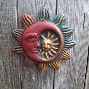 Puede incluir: Una placa de pared circular que presenta una luna creciente roja con un perfil de rostro humano que se superpone a un sol dorado más pequeño con rostro humano. El borde exterior consta de rayos verdes y dorados alternos con textura.