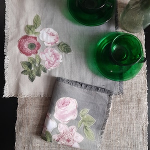 以下が含まれることがあります： 花柄の刺繍とフリンジの付いたグレーのリネン製のランチョンマット2枚。ランチョンマットは、緑色のガラス製のティーカップとソーサー2組が置かれた麻の表面の上に置かれています。