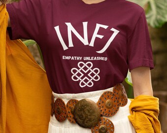 Pode incluir: Uma camiseta vinho com texto branco que diz "INFJ Empathy Unleashed" e um símbolo de infinito branco. A camiseta está sendo usada por uma pessoa com um chapéu marrom e uma saia branca.