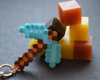 May include: A pixelated pickaxe keychain in shades of blue, brown, and orange, with a gold-colored chain. Beside it are several colorful square blocks in shades of orange, yellow, and brown.